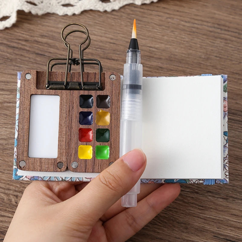 Small wooden art set with watercolor paints, brush, and paper held by a hand on a wooden surface.