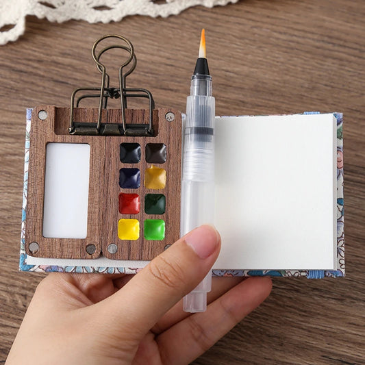 Small wooden art set with watercolor paints, brush, and paper held by a hand on a wooden surface.
