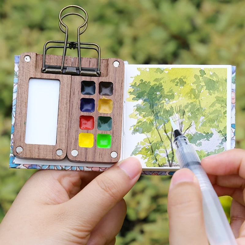 Small watercolor book with paint swatches and a tree illustration, held open by a clip with a hand holding a pen.