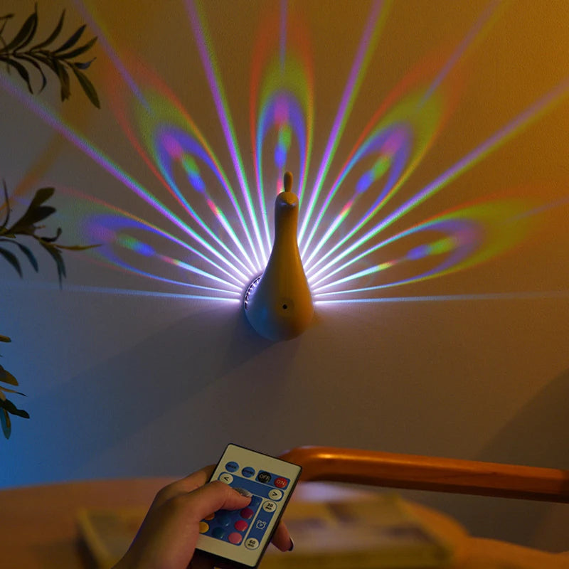 Colorful peacock night light projector on a wall with a remote control held by a hand.