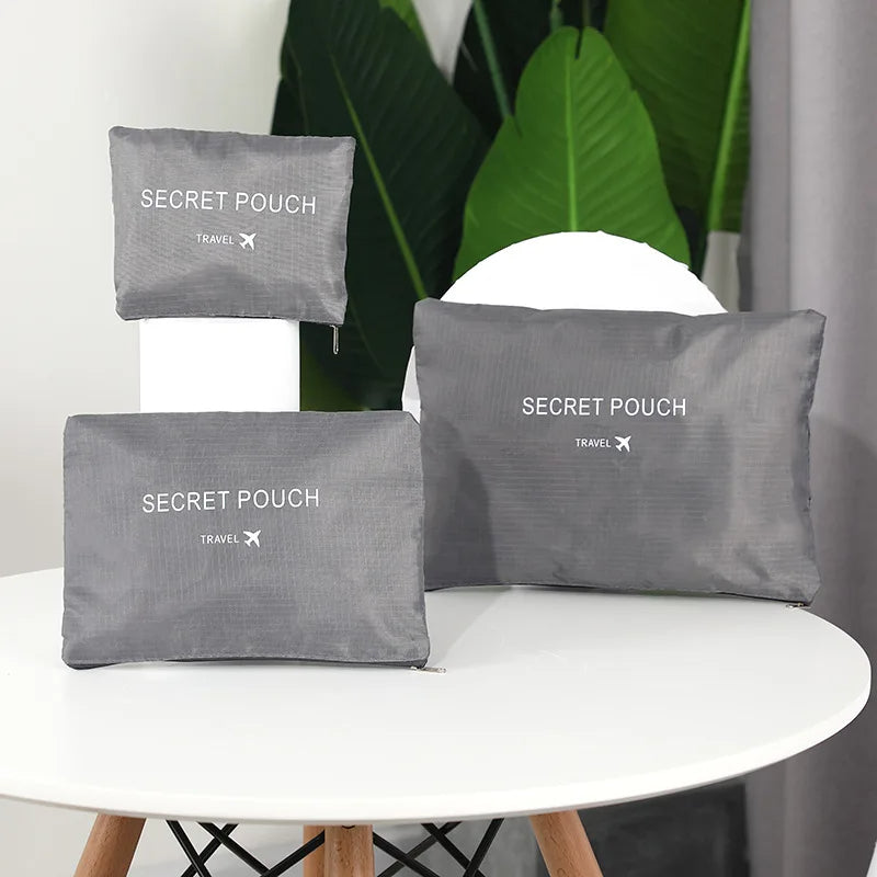 Three gray 'SECRET POUCH' travel bags on a white surface with green plants in the background.