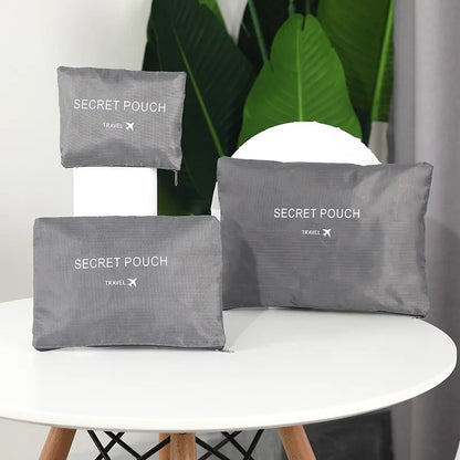 Three gray 'SECRET POUCH' travel bags on a white surface with green plants in the background.