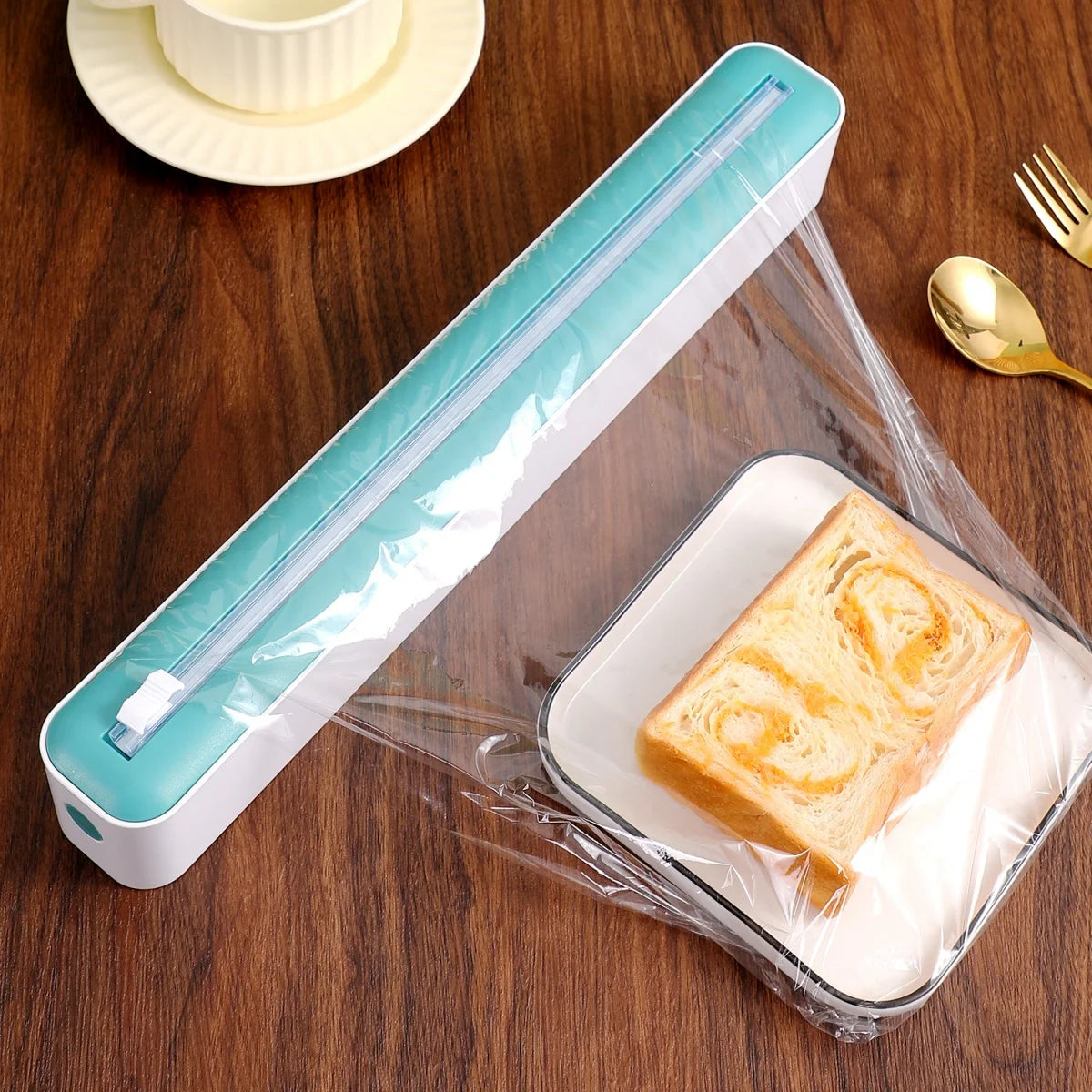 Plastic wrap dispenser with a sheet of plastic wrap covering a slice of bread on a wooden table.